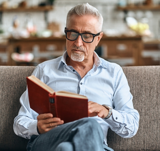 Älterer Mann mit Gleitsichtbrille sitzt auf dem Sofa und liest ein Buch.